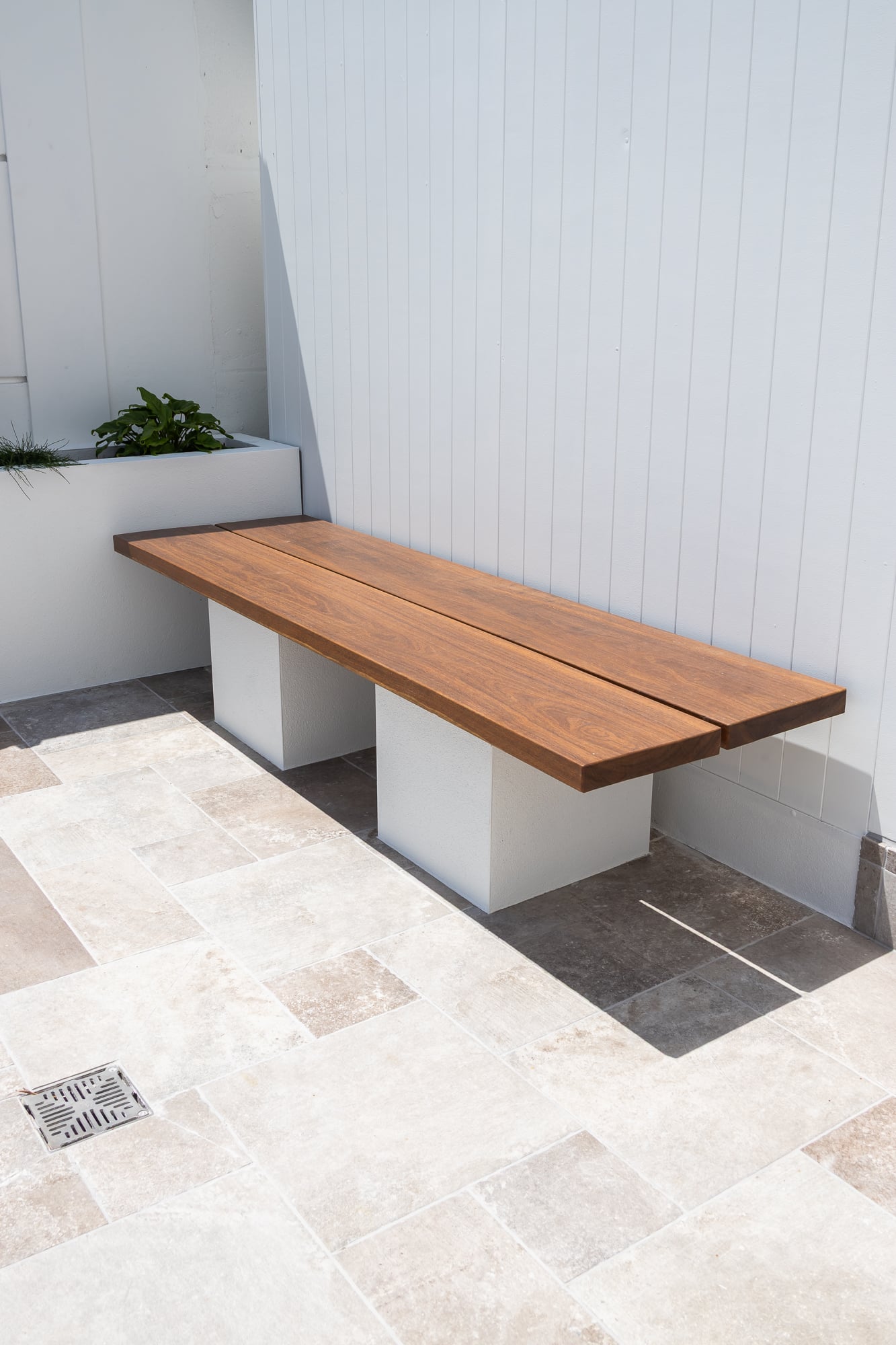 Square stainless grate drain set flush into travertine paving beside a timber bench seat