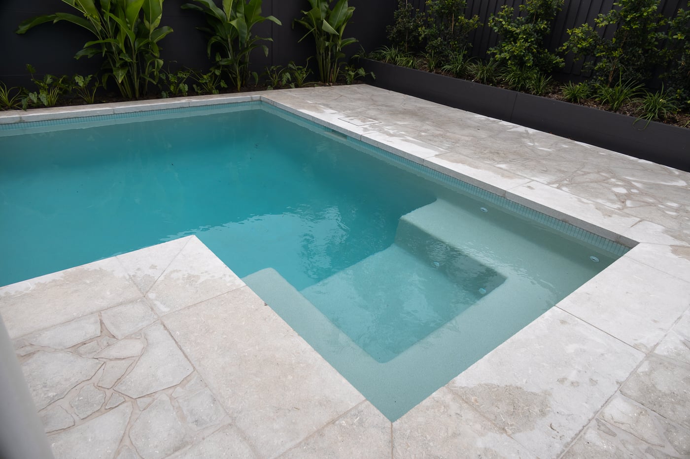 Pool with a mosaic-tiled waterline, pale stone coping and irregular flagstone paving continuing to a planted bed along a dark fence